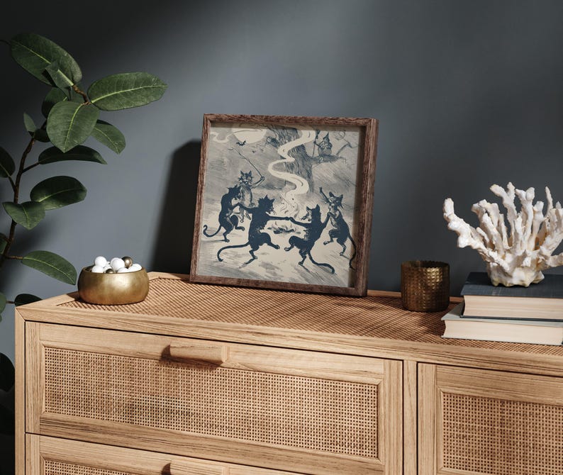 May include: A framed print featuring a vintage illustration of dancing black cats in a forest setting. The artwork is in shades of blue and gray, set in a wooden frame. The print is displayed on a light wood cabinet.