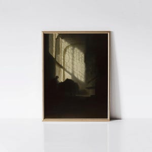 May include: A framed print of a dimly lit interior scene. The artwork features a person seated near a window with light streaming through, creating a contrast of light and shadow. The frame is gold.