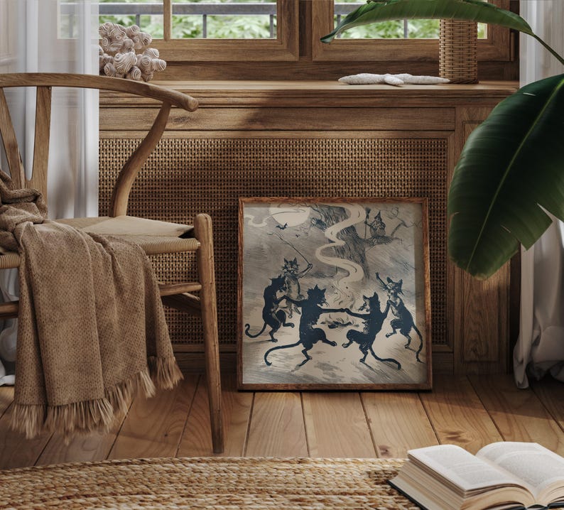 May include: A framed vintage print depicts silhouetted cats dancing in a circle under a full moon. The artwork, in shades of blue and gray, is set against a backdrop of a spooky tree and a distant house. The print is displayed on a wooden floor.