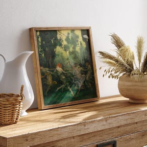 May include: A framed vintage painting with a wooden frame depicts figures dancing in a forest setting. The artwork features a palette of greens, yellows, and browns. The painting rests on a wooden dresser, next to a white pitcher and a woven basket.