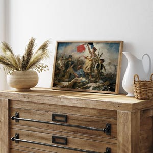 May include: A framed print of a historical painting depicting a battle scene with figures holding flags. The artwork features a woman holding a flag, with a dynamic composition and a wooden frame. The print is displayed on a wooden dresser with decorative hardware.
