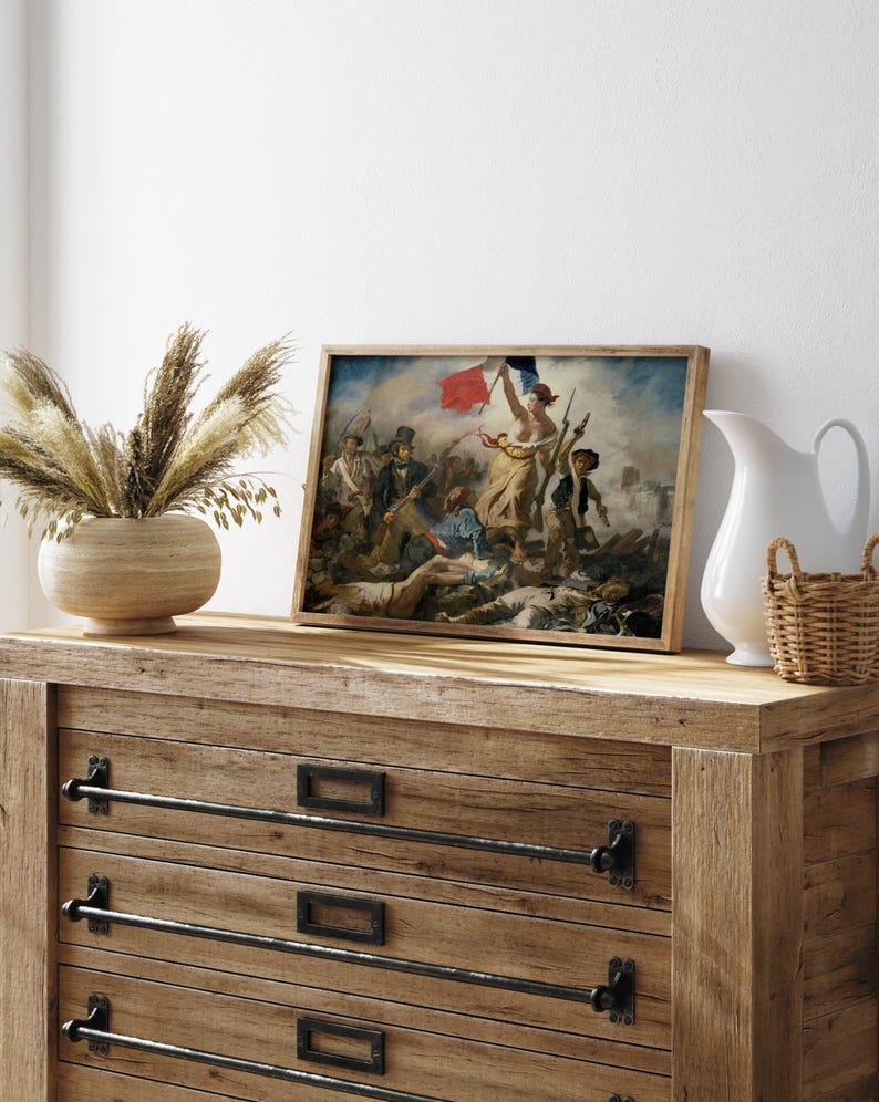 May include: A framed print of a historical painting depicting a battle scene with figures holding flags. The artwork features a woman holding a flag, with a dynamic composition and a wooden frame. The print is displayed on a wooden dresser with decorative hardware.