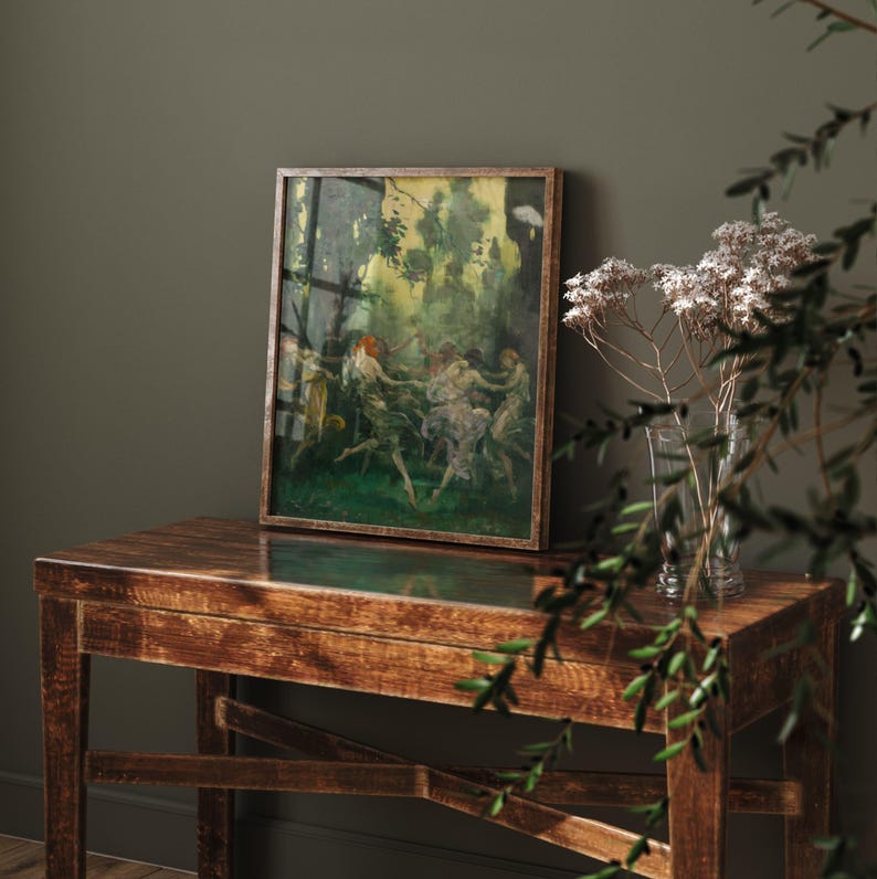 May include: A framed vintage painting depicting figures dancing in a forest setting. The artwork features a wooden frame and is displayed on a dark wooden table. A glass vase with dried flowers sits nearby, with green foliage in the foreground.