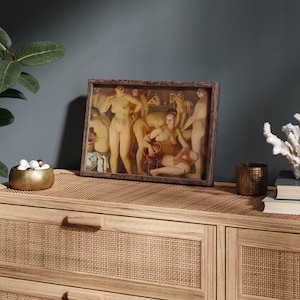 May include: A framed artwork depicting a classical scene with nude figures in warm tones. The artwork is displayed on a light wood dresser with a woven top, alongside decorative items like a gold bowl, candle holder, and coral.