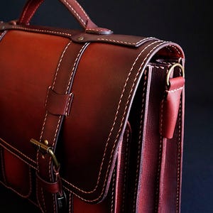 May include: A dark red leather briefcase with a top handle, two buckles, and a flap closure. The bag has a strap and a pocket on the front.