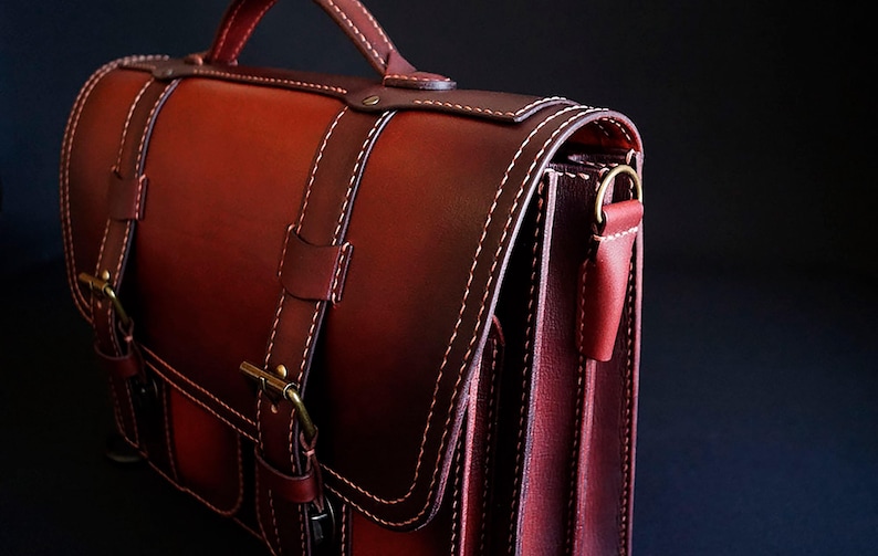 May include: A dark red leather briefcase with a top handle, two buckles, and a flap closure. The bag has a strap and a pocket on the front.