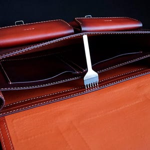 May include: A brown leather briefcase with multiple compartments and a stitching detail. The briefcase is open and a metal tool is visible inside.