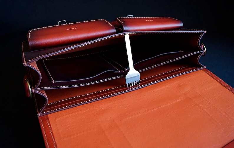 May include: A brown leather briefcase with multiple compartments and a stitching detail. The briefcase is open and a metal tool is visible inside.