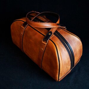 Peut inclure: Un sac de voyage en cuir marron avec des finitions noires et une fermeture &agrave; glissi&egrave;re. Le sac a une poign&eacute;e sup&eacute;rieure et une bandouli&egrave;re.