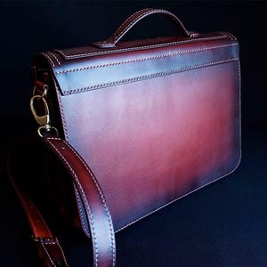 May include: A burgundy leather briefcase with a top handle and a long adjustable shoulder strap. The bag has a flap closure and a subtle distressed look.
