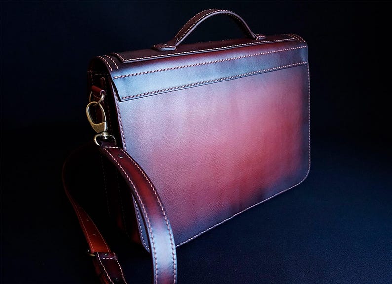 May include: A burgundy leather briefcase with a top handle and a long adjustable shoulder strap. The bag has a flap closure and a subtle distressed look.