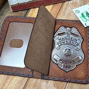May include: A brown leather badge holder with blue stitching featuring a silver-toned Manheim Township Police badge with the number 140. The holder includes a clear plastic window for identification and a protective flap over the metal badge.