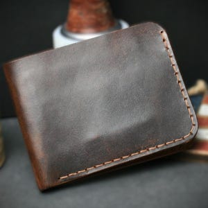 May include: A brown leather wallet with a bi-fold design. The wallet has a smooth, textured surface and is stitched with a contrasting thread. The edges are rounded, and the overall design is simple and classic.