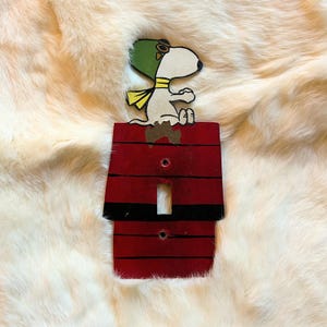 Peut inclure: Une plaque d'interrupteur rectangulaire peinte pour ressembler à une niche rouge avec des lignes horizontales noires. Un chien blanc de dessin animé avec une casquette d'aviateur verte et une écharpe jaune est assis sur le toit. Deux trous de vis.