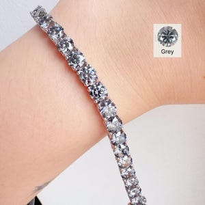 Könnte beinhalten: Ein silbernes Armband mit mehreren runden, grauen Edelsteinen. Die Edelsteine sind eng aneinander gesetzt und erzeugen eine durchgehende Linie aus Glanz. Das Armband wird an einem Handgelenk gezeigt, mit einer kleinen Grafik mit der Aufschrift "Grey".