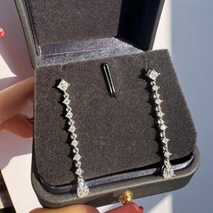 Può includere: Eleganti orecchini pendenti in argento con una serie di pietre scintillanti simili a diamanti. Ogni orecchino presenta una parte superiore quadrata, una catena di pietre più piccole e una pietra a forma di pera nella parte inferiore. Presentati in un cofanetto nero.