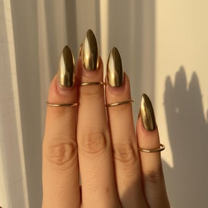 May include: Close-up of a hand with gold-colored, pointed fingernails and thin gold rings on the fingers. The nails have a shiny, metallic finish. The background is a neutral, off-white color with shadows.