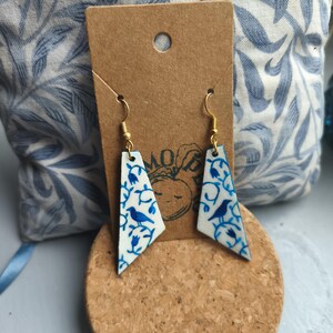 Könnte beinhalten: Weiß-blaue Ohrringe mit Muster und goldfarbenen Haken. Die Ohrringe zeigen ein Vogel- und Blumendesign. Die Ohrringe werden auf einem Korkuntersetzer und einem braunen Etikett präsentiert.