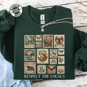 May include: Dark green sweatshirt adorned with a collage of vintage postage stamps showcasing wildlife and the phrase "RESPECT THE LOCALS." The stamps feature a deer, bear, eagle, and other animals. A silver necklace is draped across the sweatshirt.