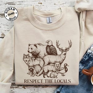 May include: A sand-coloured sweatshirt with a brown illustration of wildlife: a bear, wolf, eagle, deer, fox, raccoon, rabbit, and duck. The text "RESPECT THE LOCALS" is printed below. The Gildan brand logo is in the upper left corner.