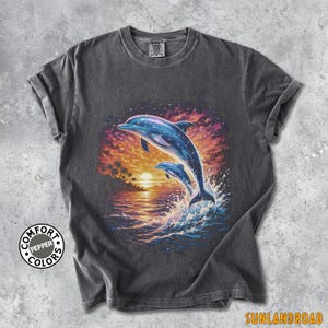 May include: Dark grey t-shirt with a colourful graphic of two blue dolphins jumping from waves, set against a sunset. The design includes palm trees and the text "SUNLANDROAD". The shirt has rolled sleeves and the text "COMFORT COLORS".