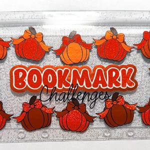 May include: A clear, glittery plastic cash envelope featuring the text BOOKMARK Challenges in orange and white. The design includes ten orange and brown pumpkins with bows, along with small autumn leaves scattered across the surface.