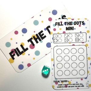 May include: Two white game cards with the text "FILL THE DOTS" and "FILL THE DOTS MINI" in black. The cards have colourful polka dots. A green die with white dots is next to the cards. Game instructions are printed at the bottom.
