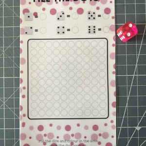 May include: A pink and white game card with the text "FILL THE DOTS" and a grid of circles. The card has a pink polka dot pattern. A translucent pink die with white dots is next to the card.