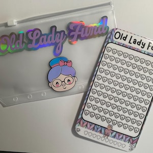 K&ouml;nnte beinhalten: Eine transparente Rei&szlig;verschlusstasche und ein Spar-Tracker mit dem Text "Old Lady Fund". Die Tasche hat ein holografisches Etikett und einen Cartoon einer Frau mit Brille. Der Spar-Tracker hat einen gemusterten Rand und ein Raster aus Schilden.