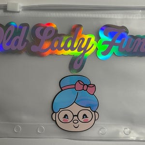 K&ouml;nnte beinhalten: Transparente Kunststofftasche mit Rei&szlig;verschluss. Der Text "Old Lady Fund" ist in holografischen, irisierenden Buchstaben geschrieben. Darunter befindet sich eine Cartoon-Illustration einer Frau mit blauen Haaren, Brille und rosa Schleife.