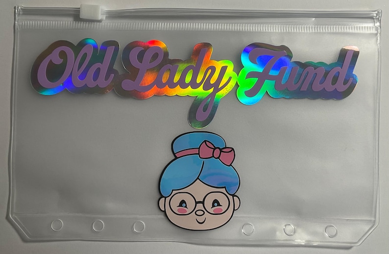 K&ouml;nnte beinhalten: Transparente Kunststofftasche mit Rei&szlig;verschluss. Der Text "Old Lady Fund" ist in holografischen, irisierenden Buchstaben geschrieben. Darunter befindet sich eine Cartoon-Illustration einer Frau mit blauen Haaren, Brille und rosa Schleife.