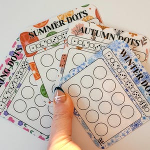 May include: Four seasonal themed cards labelled "Spring Dots", "Summer Dots", "Autumn Dots", and "Winter Dots". Each card has a different border design and a grid of circles. The cards are held by a hand.