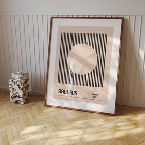 May include: A framed print with a black and white striped design and a large circle in the center. The circle is a light beige color. The print has the text "BAHAUS" in black letters and "MUNICH 1923" in smaller black letters.