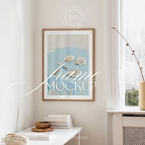 Könnte beinhalten: Ein gerahmter Kunstdruck mit einem Stillleben von Austern auf einer blauen Oberfläche. Das Kunstwerk wird in einem hellbraunen Holzrahmen präsentiert. Der Text "Frame Mockup" ist überlagert. Ein weißer Tisch trägt Bücher und eine Holzschale.