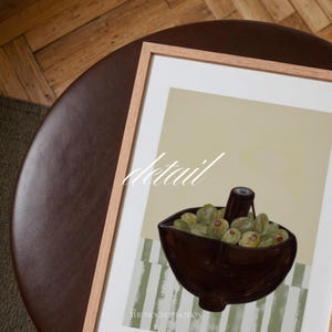 Top-down angled portrait frame mockup: light oak wood frame styled on a round dark brown leather ottoman over a textured neutral rug and parquet hardwood floor, cozy Scandinavian/minimal home-decor staging with natural light — ideal mockup for wall art prints, gallery wall, printable poster listings.