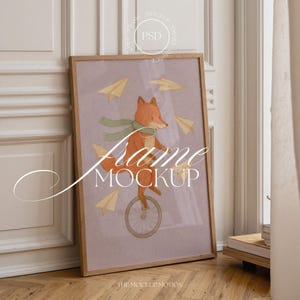Puede incluir: Una impresión artística enmarcada con una ilustración caprichosa de un zorro montando en monociclo, rodeado de aviones de papel. El zorro lleva una bufanda verde. El marco es de madera marrón claro. El texto "Frame Mockup" está en la obra.