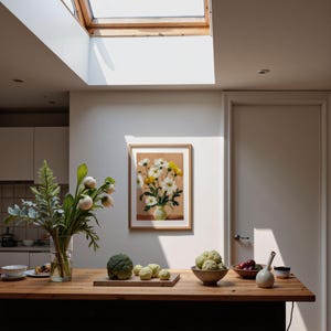 May include: A kitchen scene with a wooden countertop featuring a framed floral painting, a vase of greenery, and various fruits and vegetables. The room is illuminated by natural light from a skylight.