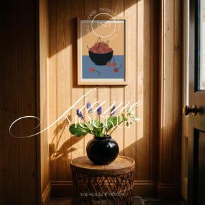 Könnte beinhalten: Ein gerahmter Kunstdruck mit einer Schale Kirschen auf blau-orangefarbenem Hintergrund hängt an einer holzgetäfelten Wand. Darunter steht eine schwarze Vase mit Blumen auf einem geflochtenen Beistelltisch. Die Szene wird von warmem Sonnenlicht durchflutet.