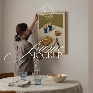 以下が含まれることがあります： トースト、卵、塩などの朝食の静物画が描かれた額入りのアートワーク。 フレームは明るい色の木材です。 テキスト「Frame Mockup」が重ねられています。 女性が壁にアートワークを掛けています。 テーブルにはグラスとボウルがセットされています。
