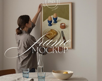 May include: A framed artwork depicting a breakfast still life with toast, eggs, and salt. The frame is light wood. The text "Frame Mockup" is overlaid. A woman is hanging the artwork on a wall. A table is set with glasses and a bowl.
