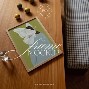May include: A framed art print featuring a butterfly design with a woman's silhouette, set on a wooden table. The frame is light brown, and the artwork has a green and blue color scheme. A glass of water and some lemons are also on the table. The text "Frame Mockup" is visible.