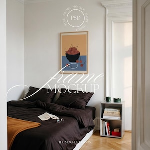 May include: A bedroom scene with a framed art print depicting cherries in a bowl. The bed has dark brown bedding and an orange throw. A small side table holds books and a green mug. The walls are white.