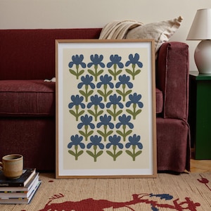 K&ouml;nnte beinhalten: Ein gerahmter Kunstdruck mit einem floralen Muster. Der Druck zeigt blaue Blumen mit gr&uuml;nen Stielen und Bl&auml;ttern auf cremefarbenem Hintergrund, in einem Holzrahmen. Das Kunstwerk wird auf einem gemusterten Teppich ausgestellt, der f&uuml;r die Inneneinrichtung geeignet ist.