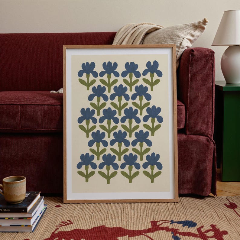 K&ouml;nnte beinhalten: Ein gerahmter Kunstdruck mit einem floralen Muster. Der Druck zeigt blaue Blumen mit gr&uuml;nen Stielen und Bl&auml;ttern auf cremefarbenem Hintergrund, in einem Holzrahmen. Das Kunstwerk wird auf einem gemusterten Teppich ausgestellt, der f&uuml;r die Inneneinrichtung geeignet ist.