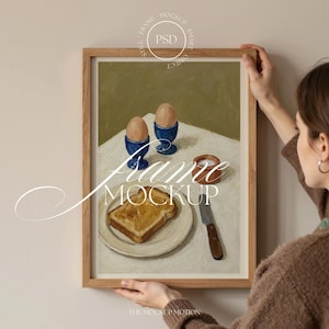 Puede incluir: Pintura enmarcada de naturaleza muerta con una escena de desayuno. La obra representa dos huevos en hueveras azules, una rebanada de tostada en un plato, un pequeño cuenco de sal y un cuchillo. El marco es de madera clara.