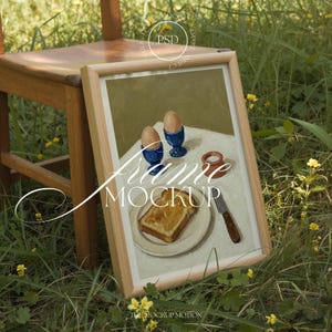 Könnte beinhalten: Gerahmtes Gemälde einer Frühstücksszene mit zwei Eiern in blauen Eierbechern, Toast und einem kleinen Salzstreuer. Der Rahmen ist aus hellem Holz, und der Text "Frame Mockup" ist sichtbar. Die Szene spielt sich im Freien ab.