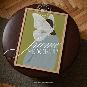 Puede incluir: Una impresión artística enmarcada con un diseño de mariposa y la silueta de una mujer. La obra de arte tiene un marco de madera clara y un fondo verde. Las palabras "Frame Mockup" se muestran. La impresión está sobre una mesa marrón.