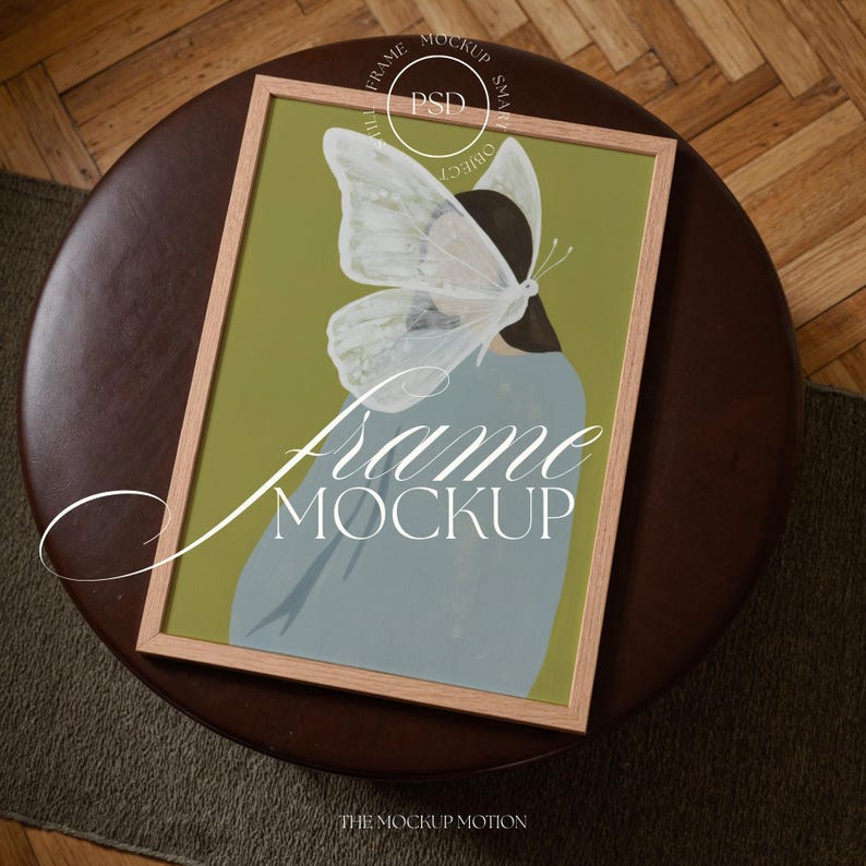 Top-down angled portrait frame mockup: light oak wood frame styled on a round dark brown leather ottoman over a textured neutral rug and parquet hardwood floor, cozy Scandinavian/minimal home-decor staging with natural light — ideal mockup for wall art prints, gallery wall, printable poster listings.