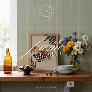 Puede incluir: Una naturaleza muerta con una impresión enmarcada, un jarrón de flores y una botella. La impresión artística presenta un patrón de frutas y cuadros. Un cuenco de higos y una pila de platos también están sobre la superficie de madera. El texto "Frame Mockup" es visible.