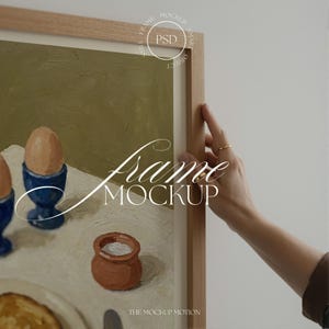 以下が含まれることがあります： 青いエッグカップに入った卵、小さな茶色のポット、トーストのスライスを描いた静物画の額装されたアートワーク。 フレームは明るい色の木材です。 テキスト「Frame Mockup」が画像に重ねられています。